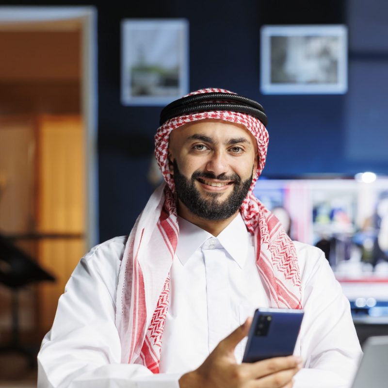 Smiling Muslim guy holding a cell phone Male Arab person in profile, grinning at camera with his personal computer close by. Using contemporary technology, smiling Muslim guy with a cell phone, portrayed in portrait with laptop on table.