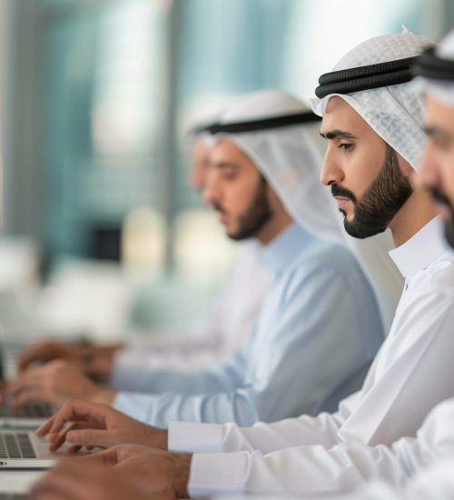Men Working on Laptops in Office A row of men wearing traditional white robes work on their laptops in a modern office setting.